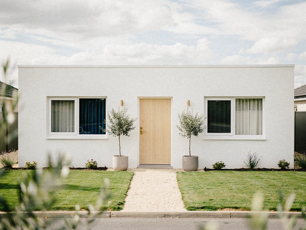 A tidy single-storey white rental home with an oak front door, olive trees and a gravel path
